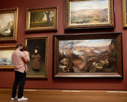 adult wearing a mask holding a small child in their arms, in front of a dark red-painted museum wall covered with ornately framed paintings of landscapes and portraits. The two are admiring a painting of a small child in historical clothes.