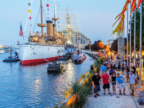 photo of waterfront walkway in late evening, many people in shorts and short-sleeve shirts playing games and standing around. In the Delaware River is docked a large old ship with smokestacks and an American flag.