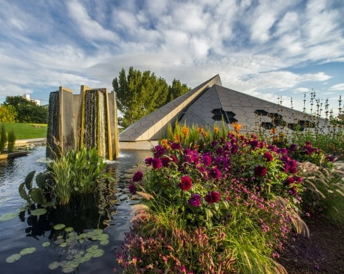various colorful plants next to a pond with a modern-art fountain in it, in front of a pyramid-shaped building