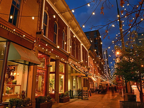 a street front at night with shop windows illuminated warmly from inside, while strings of lights reach over the sidewalk outside.